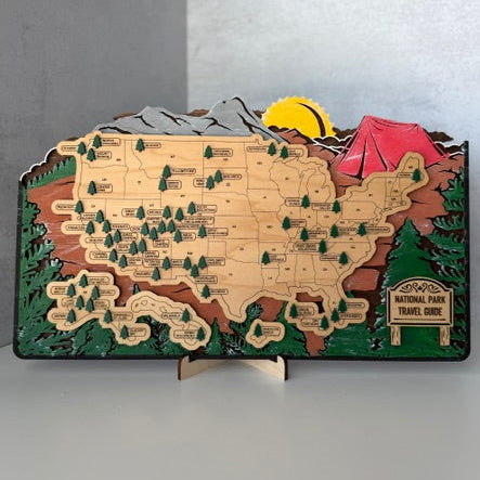 A 3D wooden map of the United States showcasing National Parks, with detailed terrain relief, a yellow sun, a red tent, and a small sign for National Park Travel Guide, displayed on a small wooden stand.