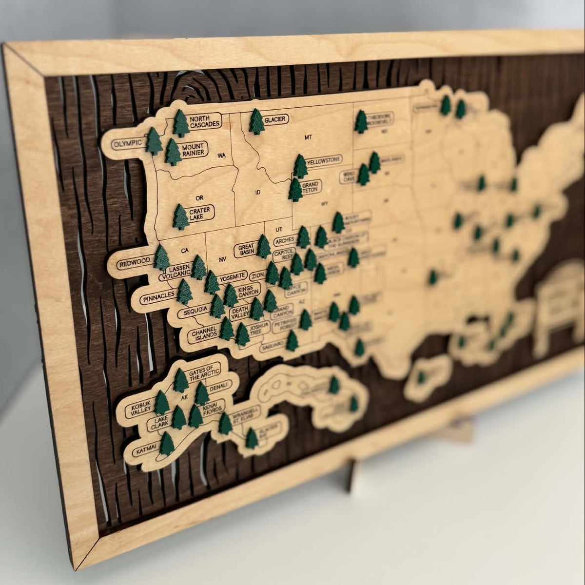 A wooden map plaque of the United States, featuring a dark wood grain background, light natural wood borders, and small green pine tree icons marking locations, displayed on a small stand against a gray wall.