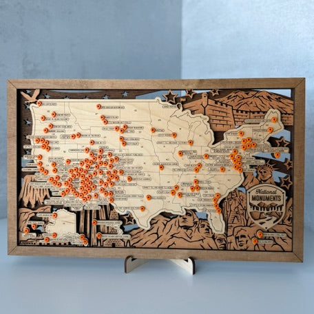 A rectangular wooden map plaque of the United States with engraved terrain details and many small orange pins scattered across it, displayed on a small stand against a light gray background.