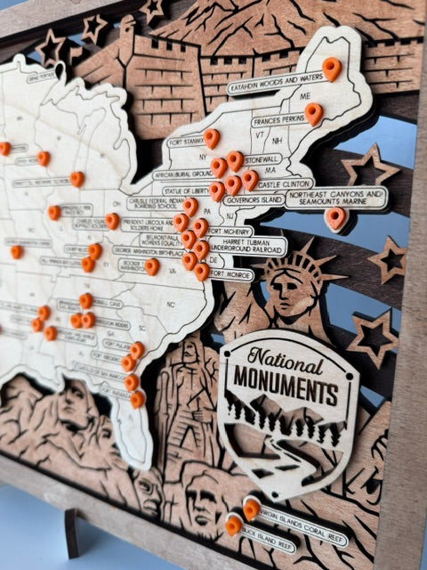 A close-up of a decorative wooden map plaque featuring labeled US monuments and orange push-pin markers, with a carved Statue of Liberty and 'National Monuments' emblem.