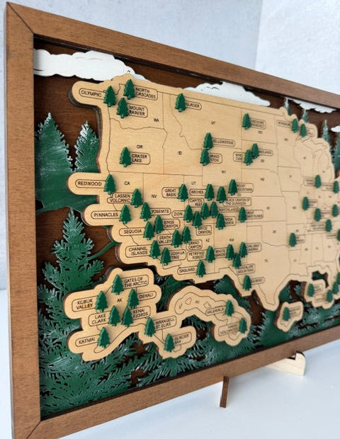 A framed wooden map wall art piece featuring the United States with labeled states, small green pine tree icons, and layered forest scenery inside a wood frame.