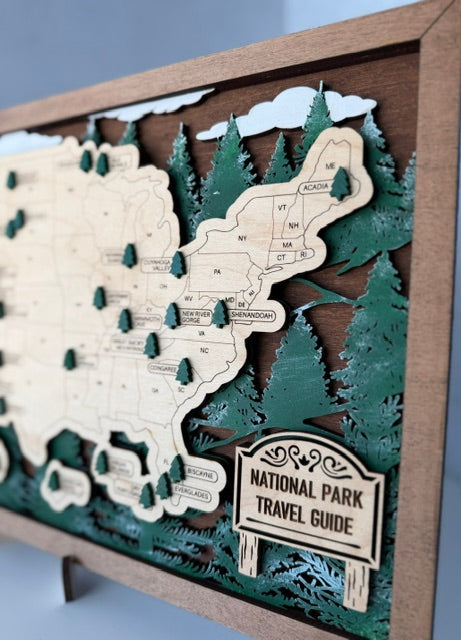 A wooden layered map of the United States styled as a National Park travel guide, featuring laser-cut pine trees and a sign, displayed on a small stand.