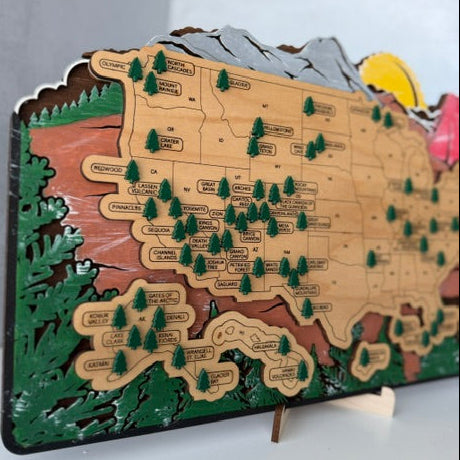 A 3D wooden USA map puzzle board with labeled states, green pine trees, mountains, forests, and a yellow sun, displayed on a white surface against a light gray wall.