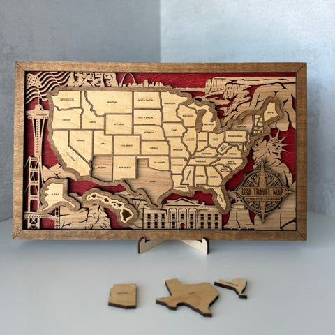 A wooden USA travel map puzzle board with engraved state outlines and landmarks, displayed on a small stand with a few loose wooden pieces in front, set against a light gray background.