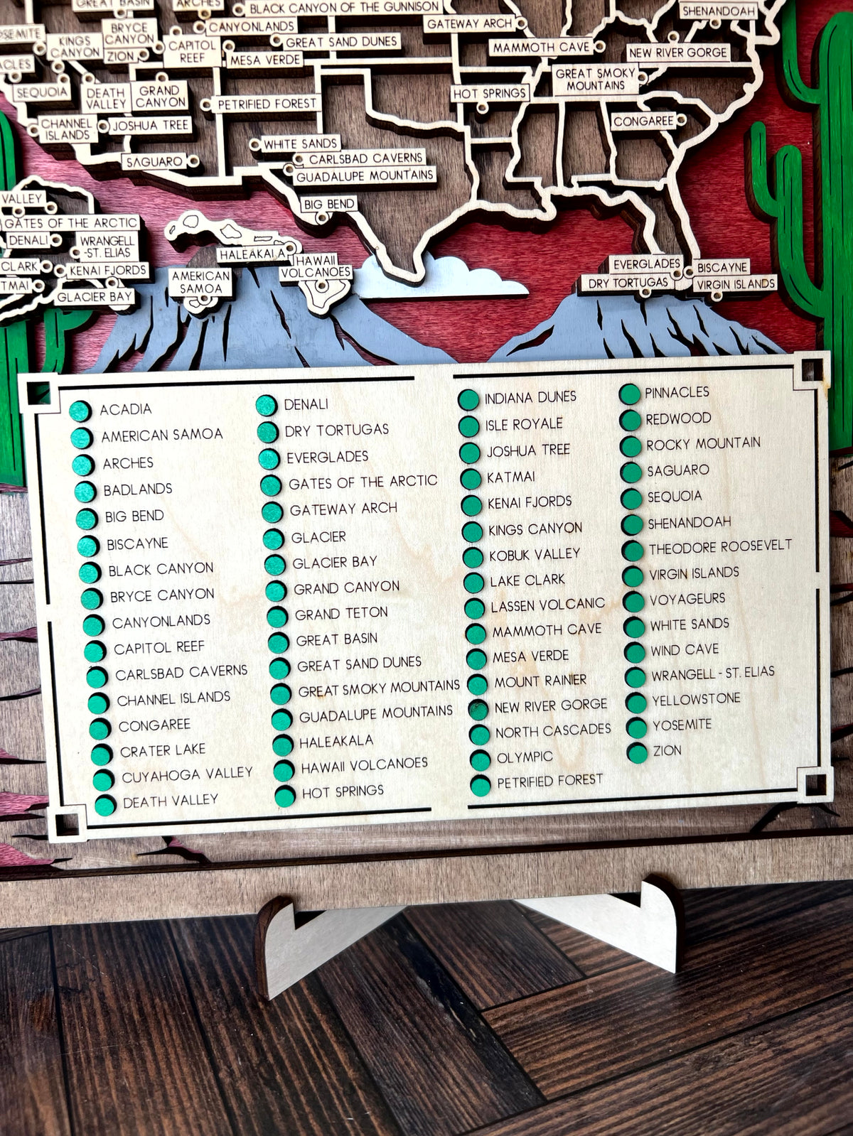 A wooden map of Arizona featuring labeled mountain peaks, desert formations, and a large white sign in front listing many trail names with colored dots, photographed on a dark wood surface.