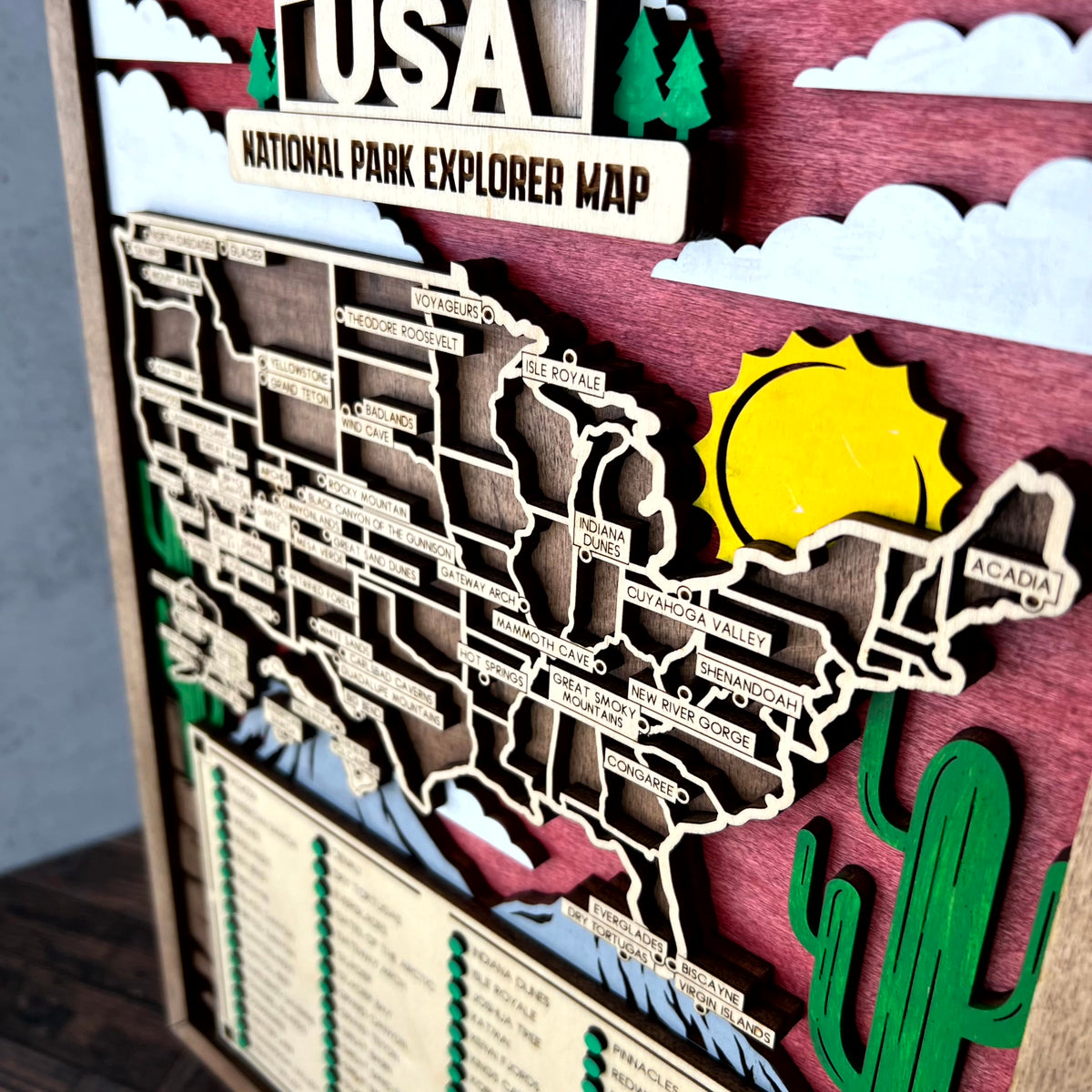 A 3D layered wooden USA National Park Explorer Map wall art piece, featuring cutout states, a yellow sun, green cactus, and a trail map legend on a dark wood background.