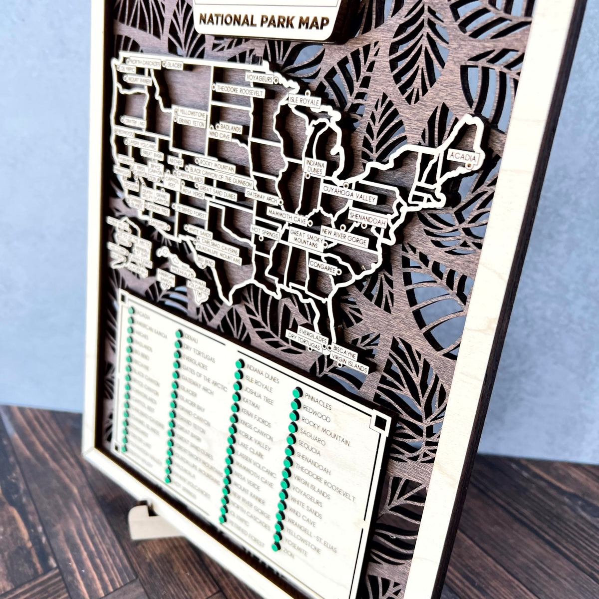 A wooden National Park Map wall sign featuring layered laser-cut USA map shapes and tropical leaf cutouts, with a list of national parks and green check markers at the bottom, shown standing on a small wooden easel against a blue wall and wood floor.