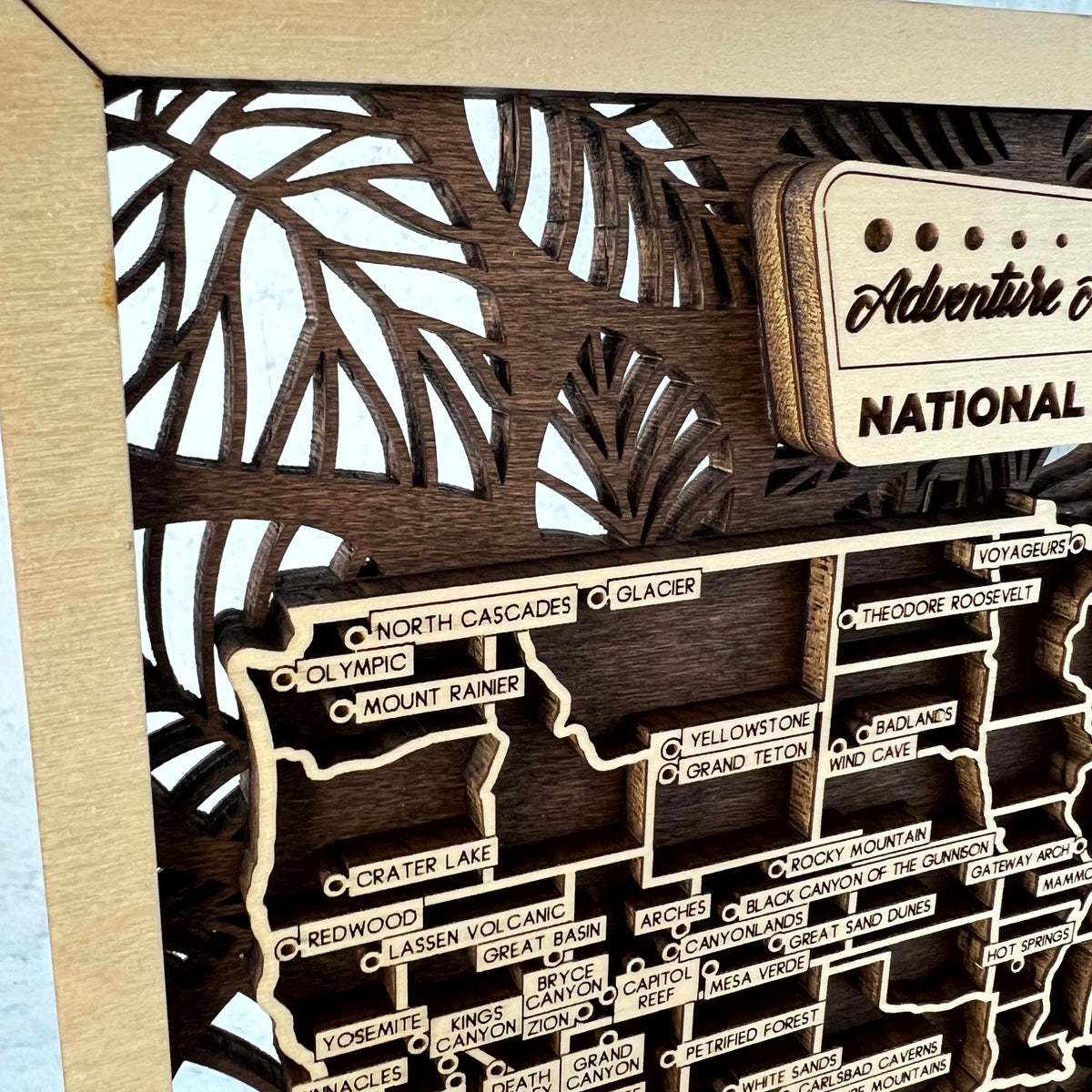 Close-up of a wooden map wall art piece featuring labeled states and scenic routes, with a decorative sign reading 'Adventure National' on a light wood background.