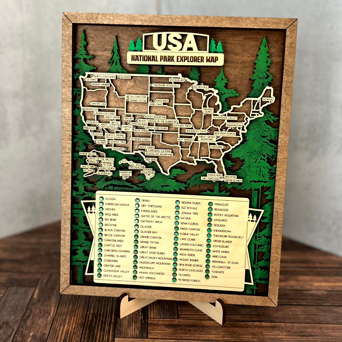 A framed wooden USA National Park Explorer Map display on a small easel stand, set on a dark wood tabletop against a light gray wall.