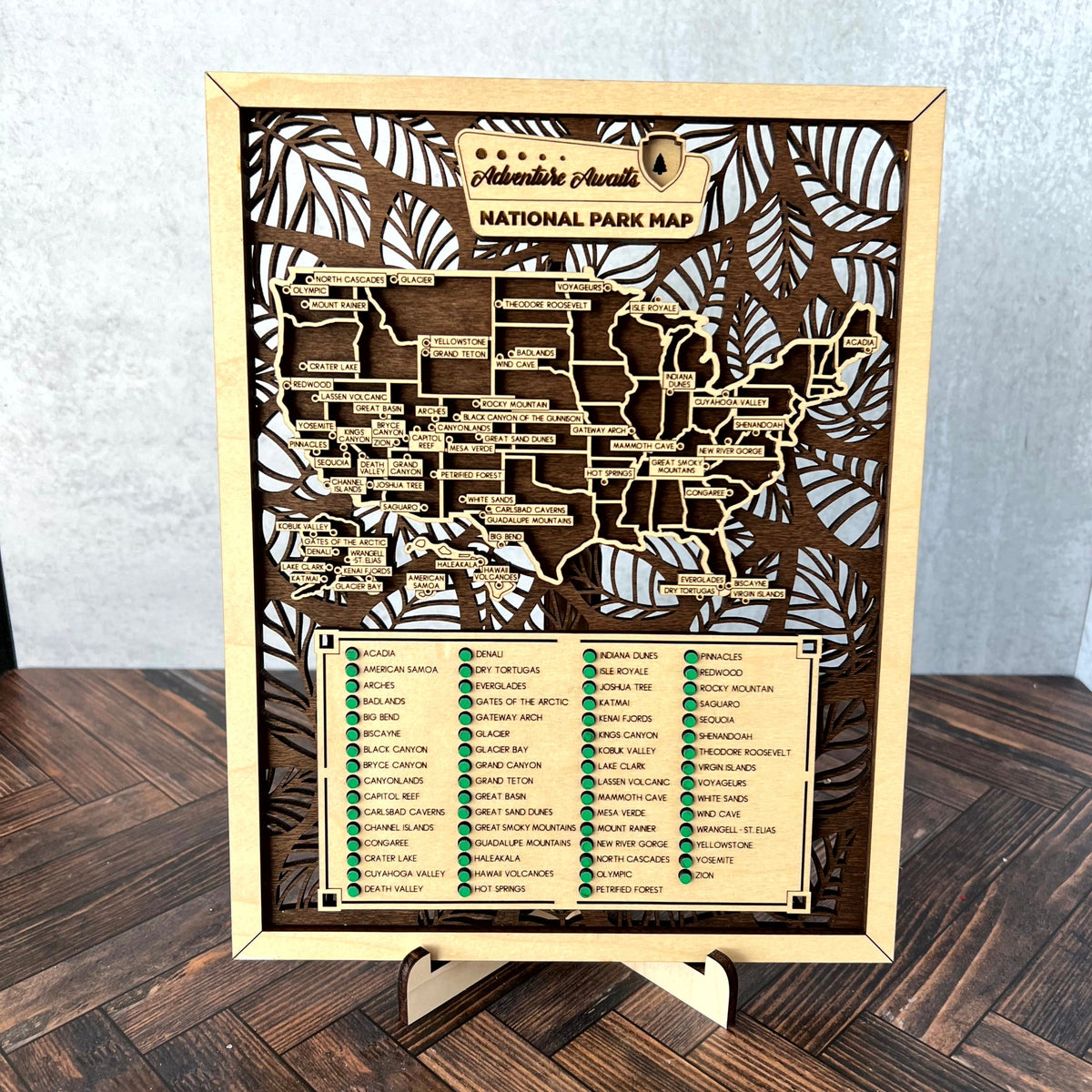 A wooden National Park Map plaque for Hawaii, featuring a laser-cut map and list of parks, displayed on a small wooden stand on a dark wood floor against a light gray wall.