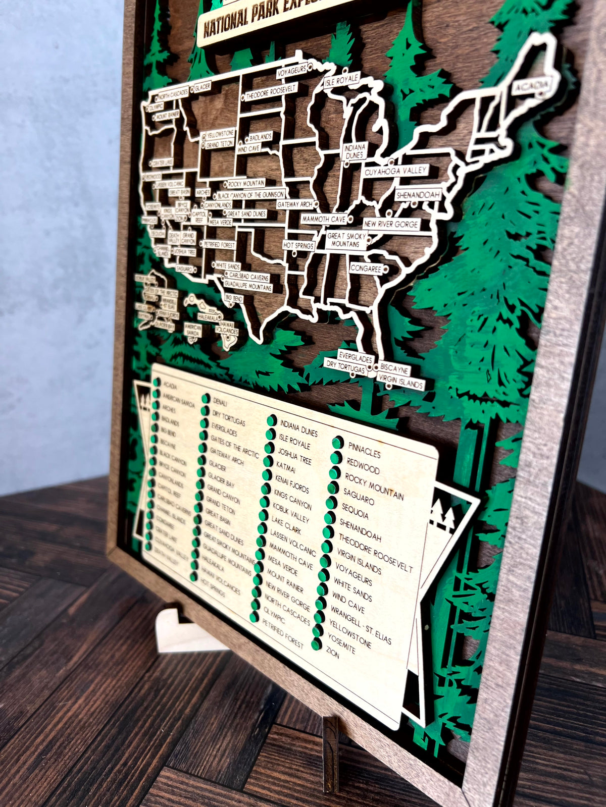 A wooden National Park Explorer map of the United States on an easel, featuring green pine trees, a list of parks, and a light blue textured wall in the background.