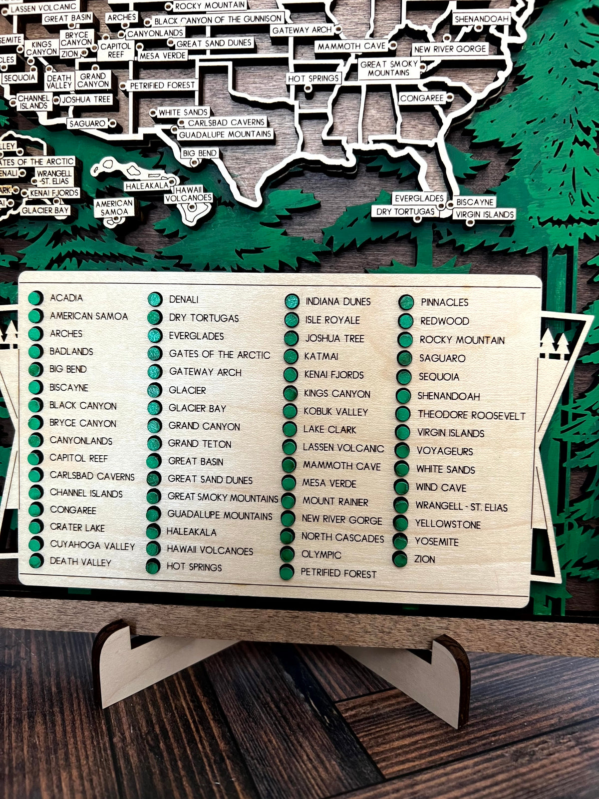 A close-up of a wooden map of Alaska with labeled locations and a large white name plaque below, placed on a dark wooden surface.