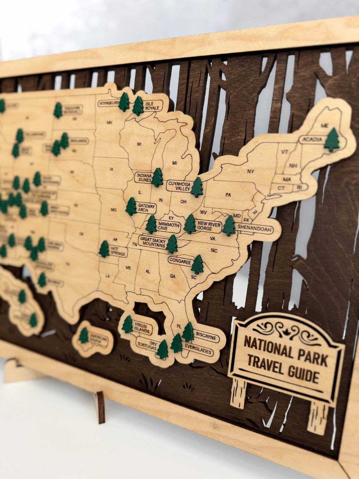 A wooden USA map wall plaque featuring a National Park Travel Guide design, with small green pine tree markers and a sign in the lower right corner, displayed on a stand.