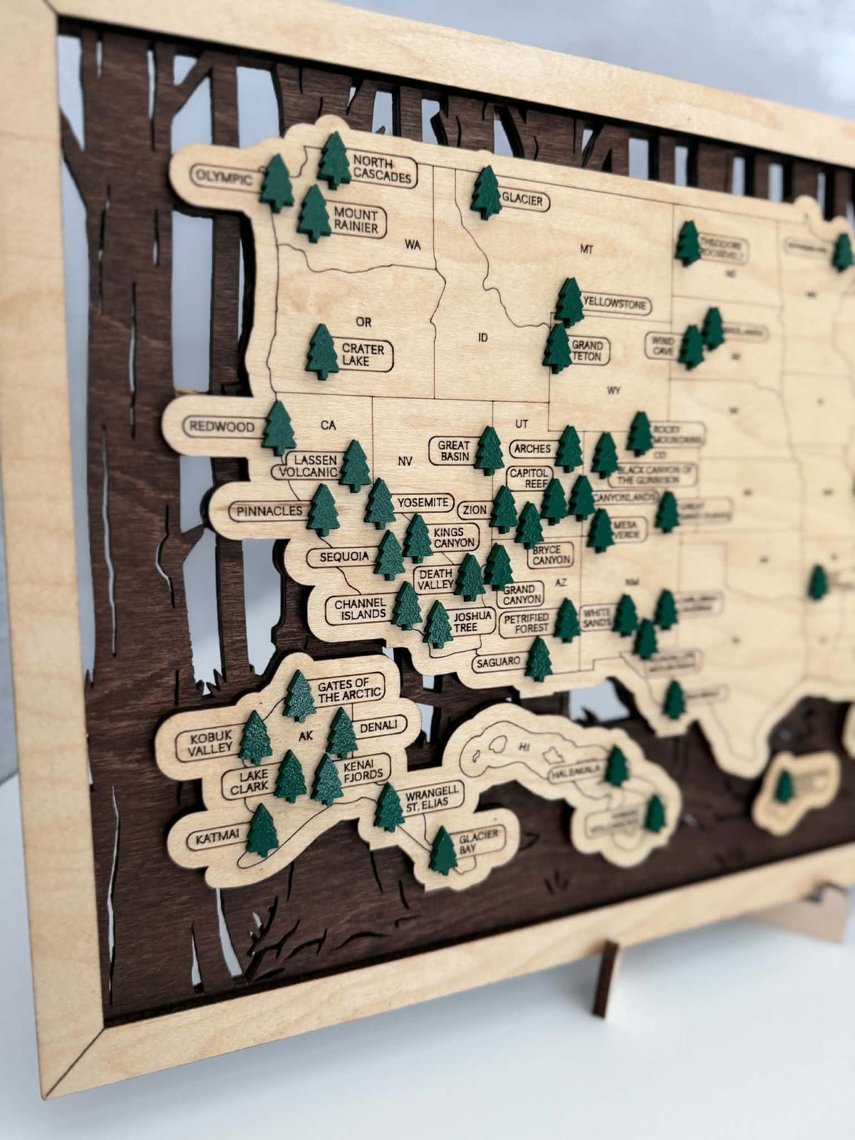 A wooden map of the United States with engraved state outlines, labeled cities, and small green pine tree markers, displayed on a stand against a light neutral background.