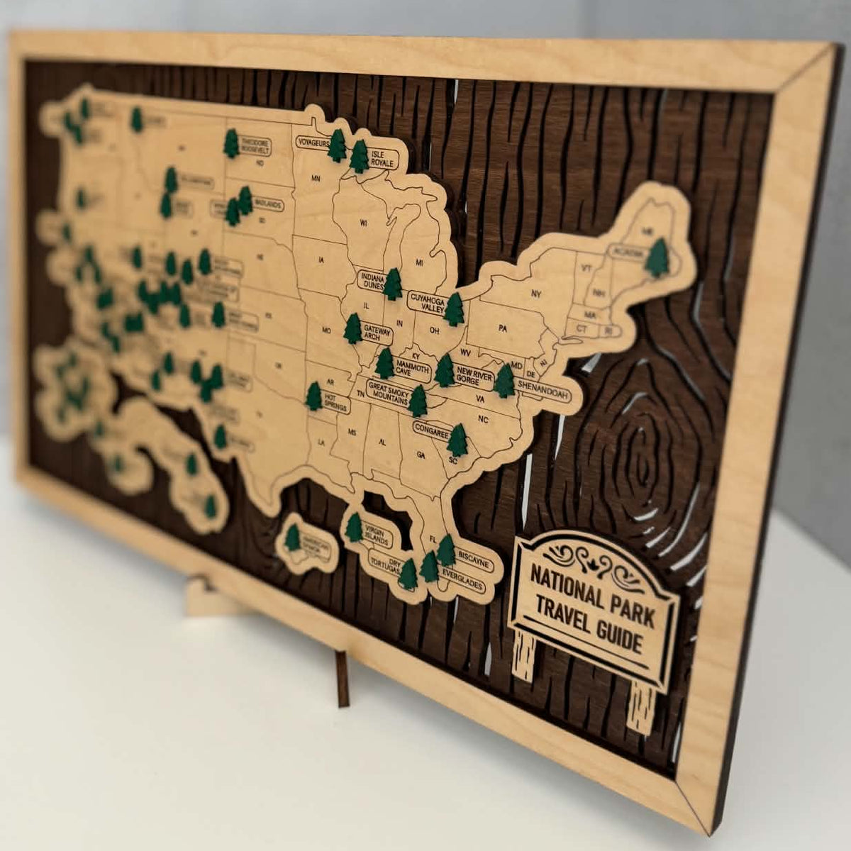 A wooden National Park Travel Guide plaque featuring an engraved map of the United States with green tree markers and a sign reading 'National Park Travel Guide,' displayed on a small stand against a light gray wall and white surface.