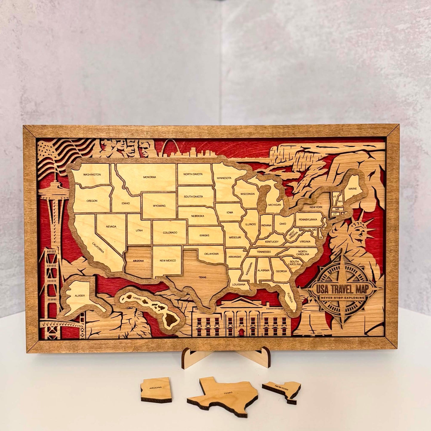 A rectangular wooden USA travel map plaque with layered laser-cut states, red background details, and small wooden puzzle pieces placed in front, photographed on a white surface against a light gray wall.