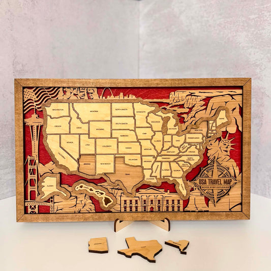 A rectangular wooden USA travel map plaque with layered laser-cut states, red background details, and small wooden puzzle pieces placed in front, photographed on a white surface against a light gray wall.